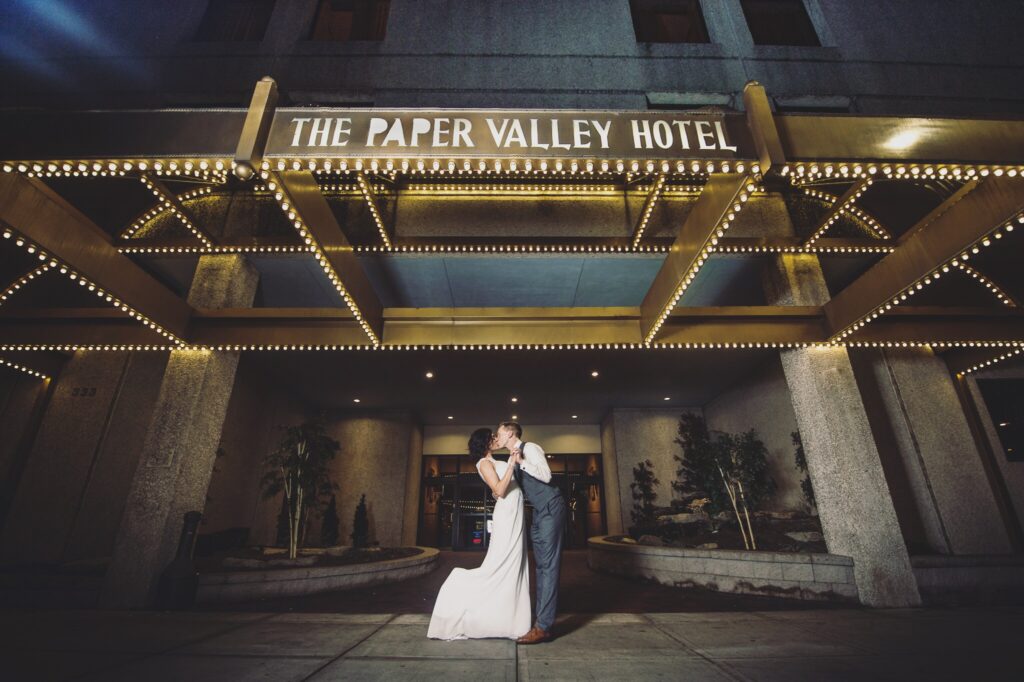 Couple in front of Paper Valley Hotel in fox valley on their wedding day kissing.
